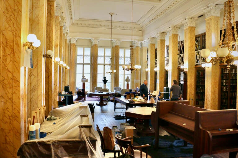 Workers make renovations in an ornate room with plastic draped over some of the desks.