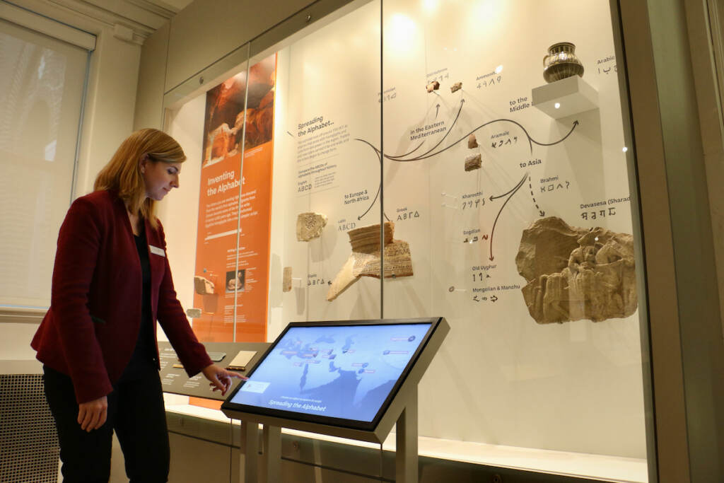 The curator stands next to a screen in front of an exhibit behind glass.