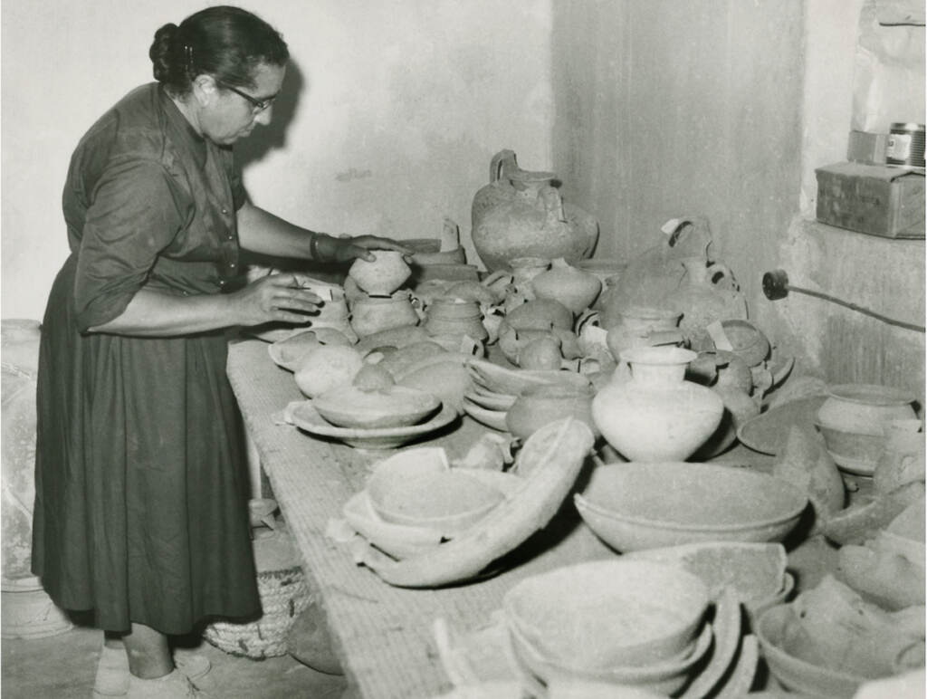 A black-and-white photo of a woman in front of a table full of artifacts.