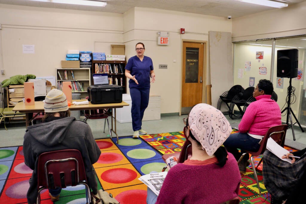 A nurse stands at the front of a room in scrubs, speaking to a class.