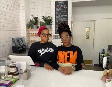 Two people stand at the counter of a store. Plants are visible in the background.