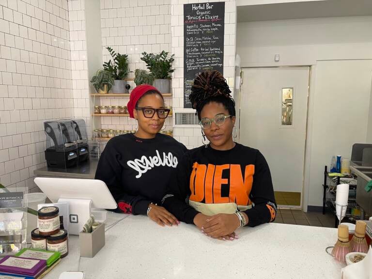 Two people stand at the counter of a store. Plants are visible in the background.