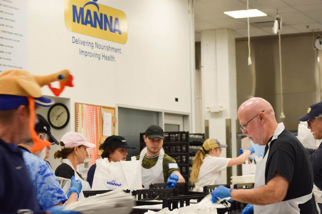 People pack up meals in a large room. A sign reads MANNA above.