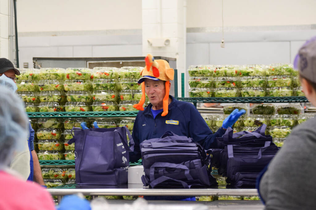 A man wearing a turkey hat stands in front of rows of packaged salads.