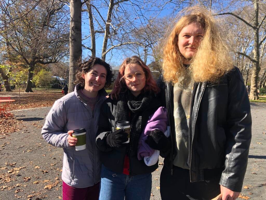 Three people stand together. A park is visible behind them.