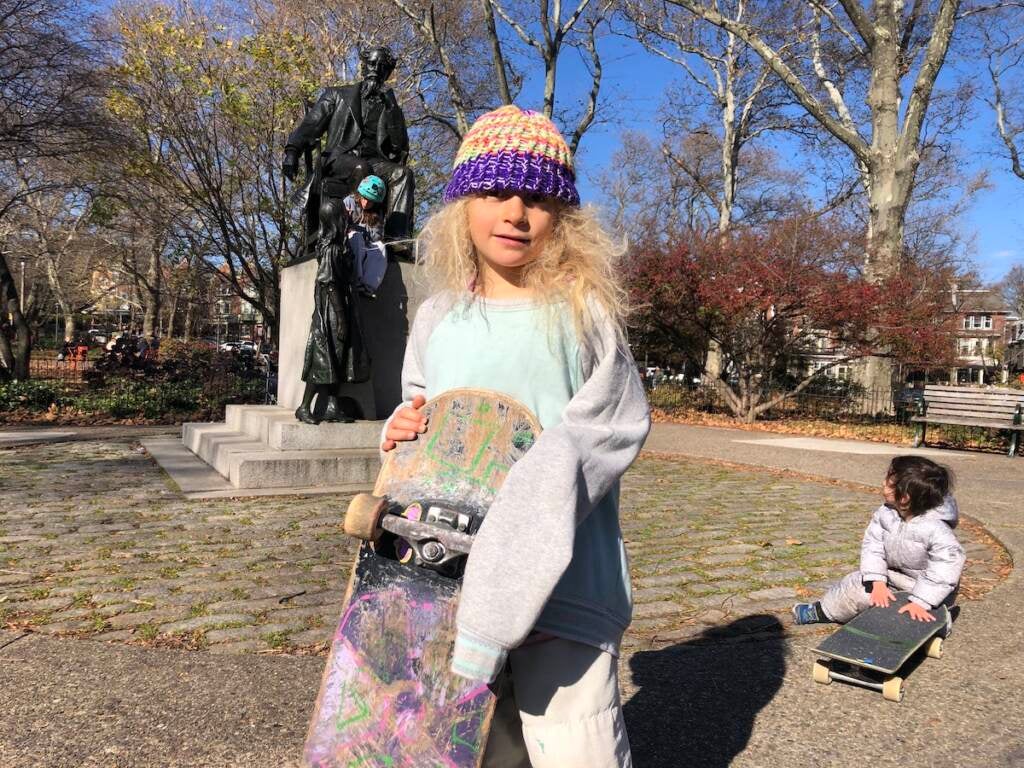 A kid poses with her skateboard. A park is visible behind her.