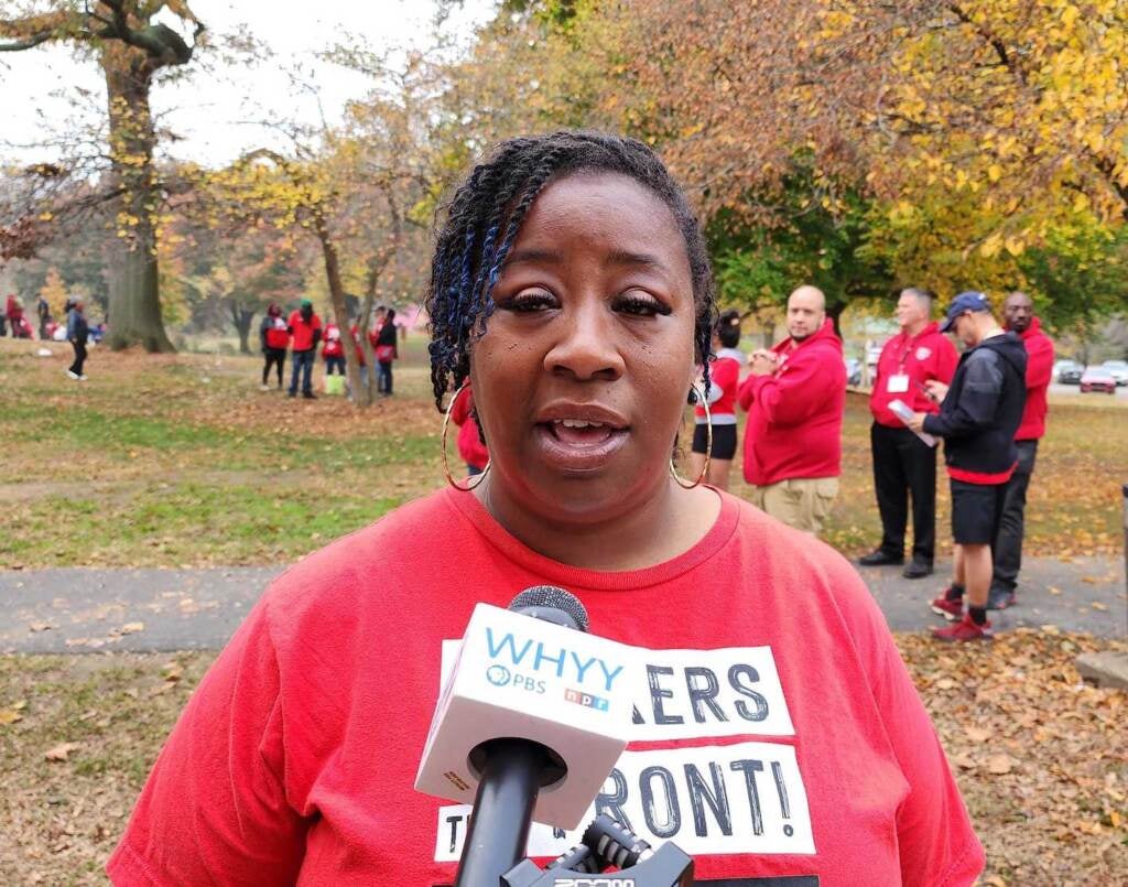 A woman speaks into a microphone, with woods visible behind her.