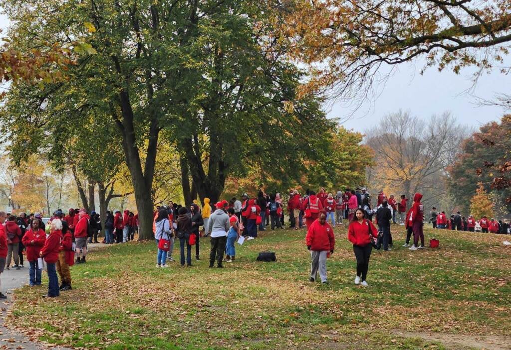 People, many wearing red T-shirts, gather in a park on a fall day.