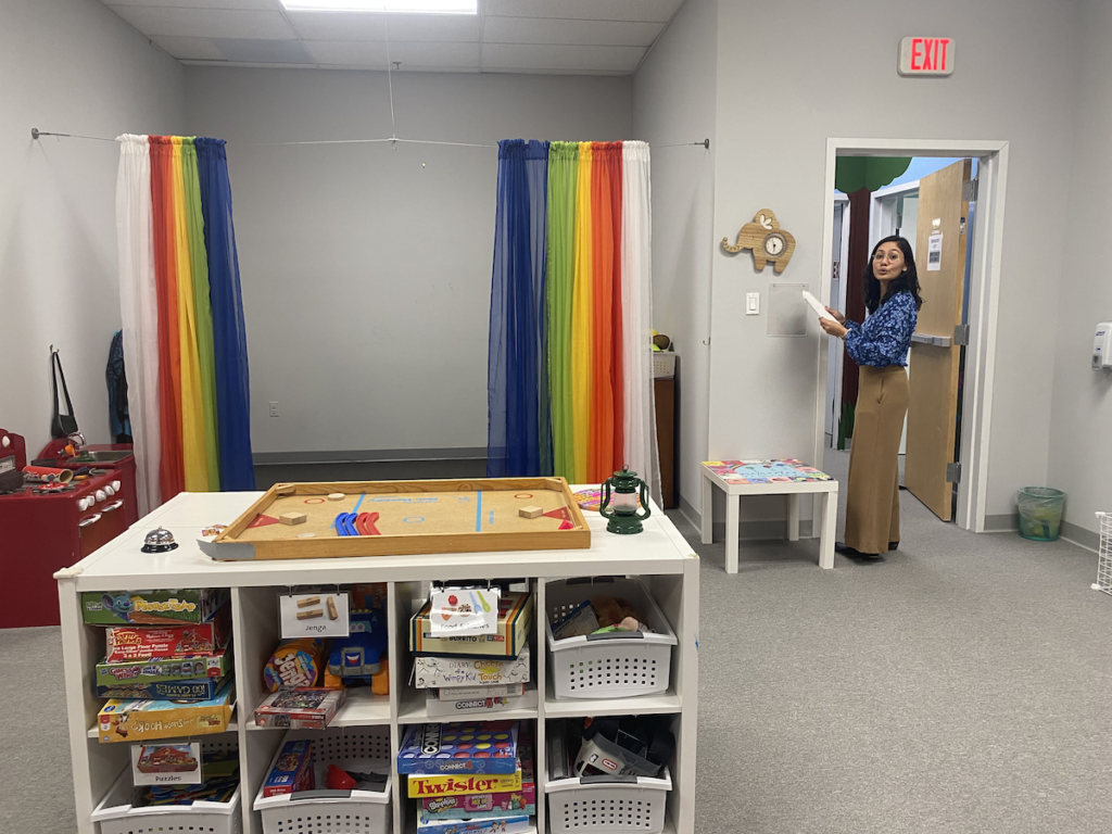 A woman stands at the doorway fo a room with a table and rainbow-colored curtains.