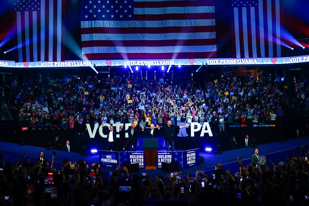 Shapiro, Obama, Biden, and Fetterman hold hands in front of a cheering crowd with an American flag displayed in the background.