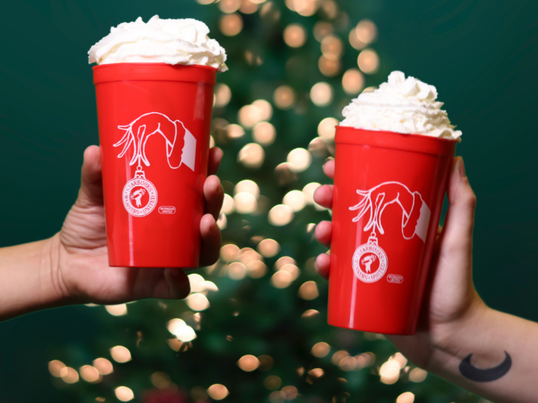 Two hands hold up red cups topped with whipped cream. A Christmas tree is visible in the background.