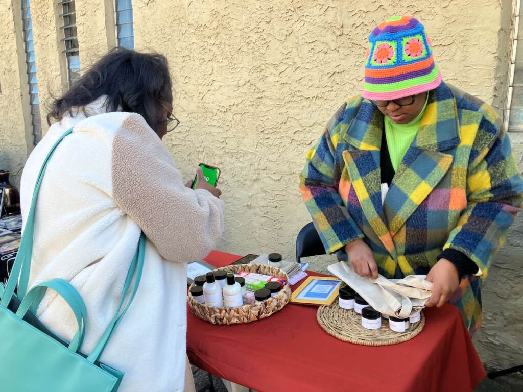 A woman arranges her products at a table as another person examines the items for sale.