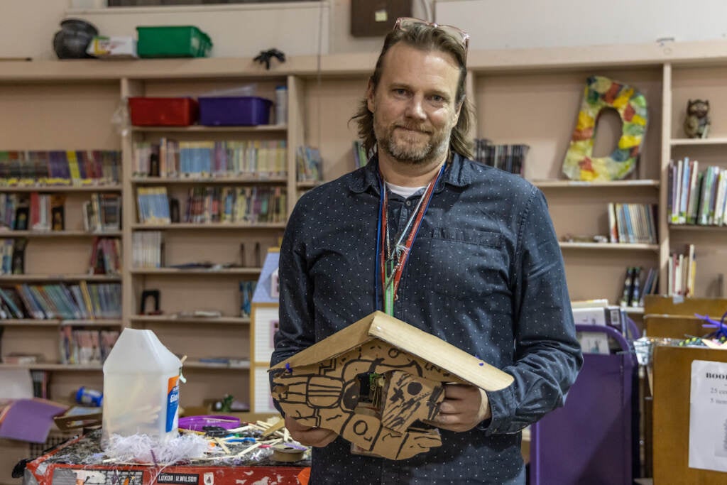 A man holds a wooden construction. Library shelves are visible behind him.