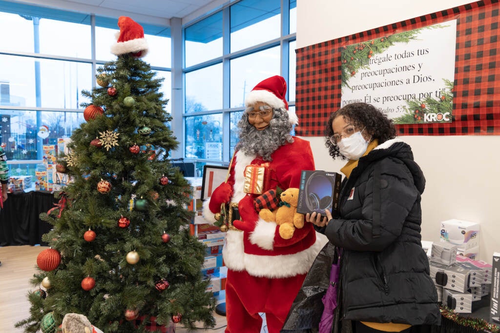 A woman holds up a box of headphones next to a person dressed as Santa and a Christmas tree.