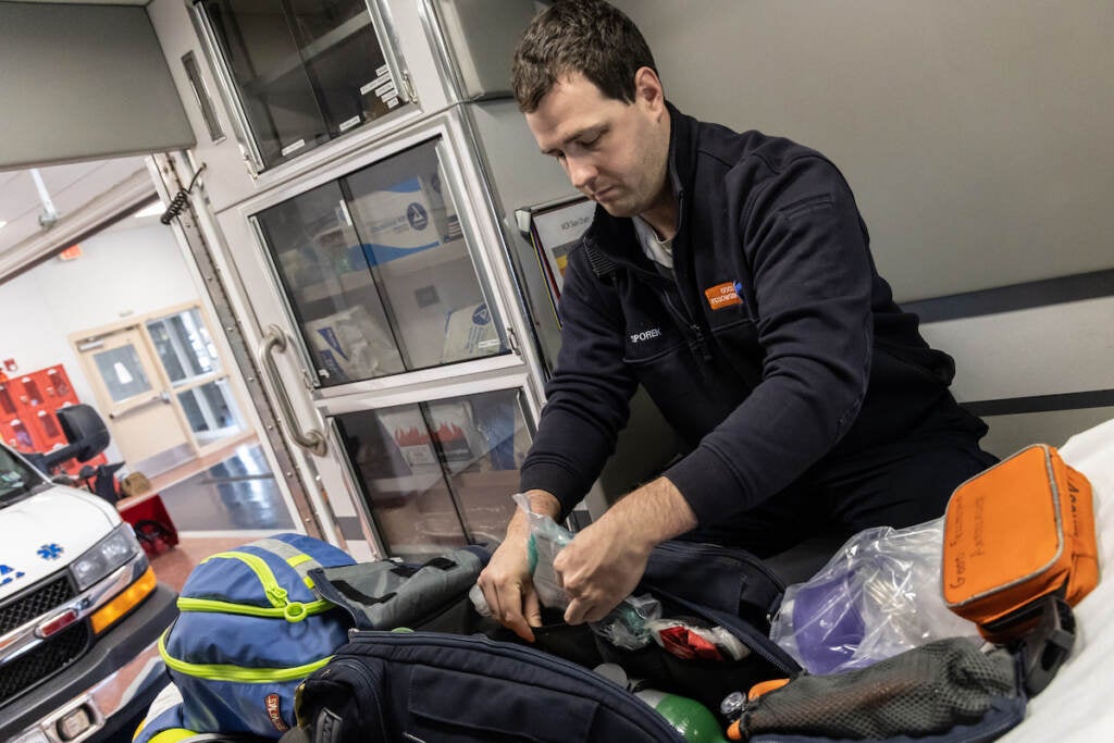 A man packs things inside of a duffel bag. An ambulance is visible in the background.