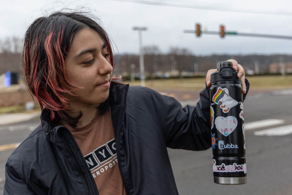 A person holds up a water bottle that has a Pride flag displayed on it. In the background, an intersection is visible.