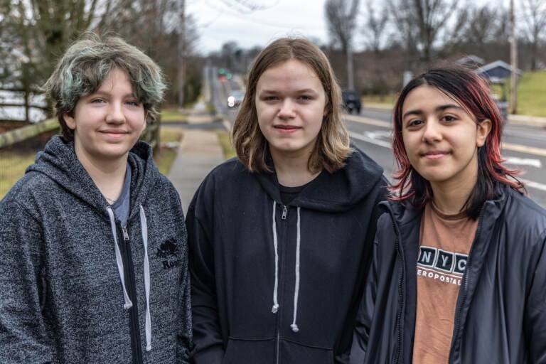 Three people stand shoulder-to-shoulder and pose for a photo. A road is visible in the background.