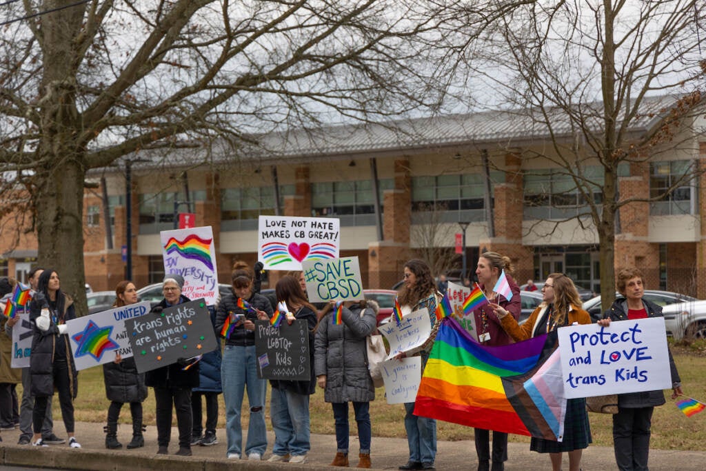 A group of protesters display the Pride flag and other signs outside of a school building.