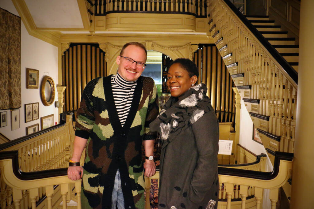 Two people pose for a photo and smile next to a staircase.