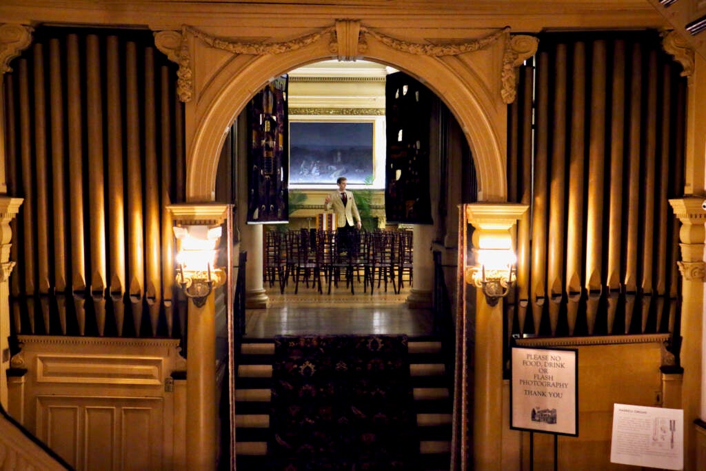 A man stands in the middle of a room visible from a staircase from across the room.