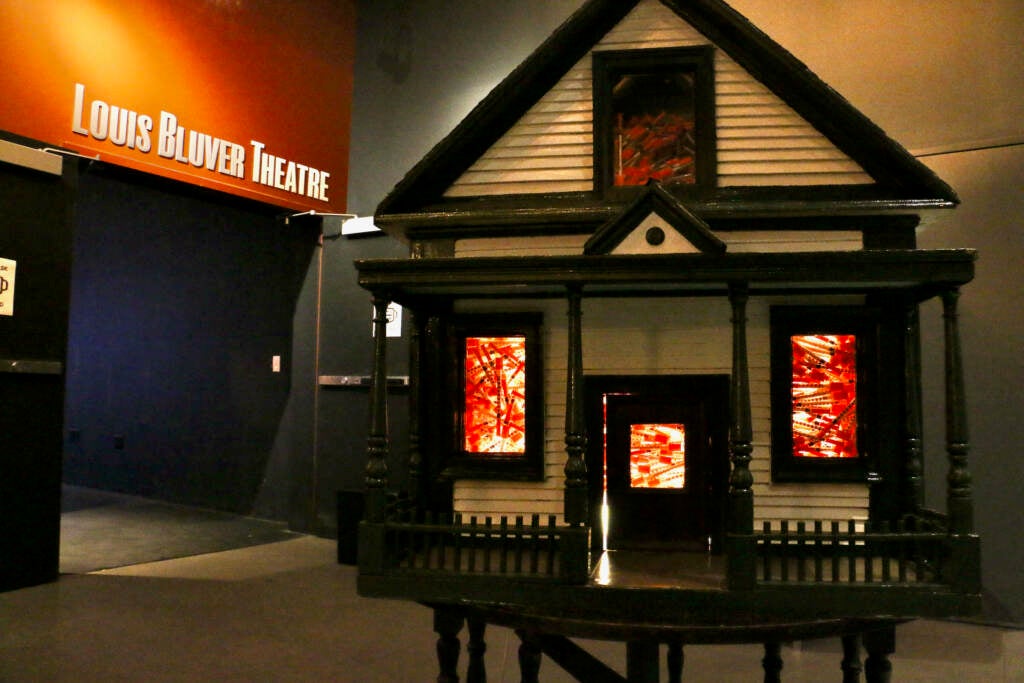 A structure of a home is lit up in front of a sign above an entrance to a hallway that reads, "Louis Bluver Theatre".