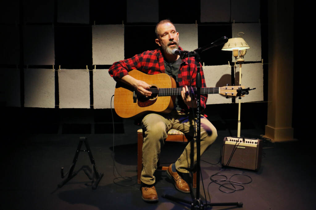 A person plays guitar into a microphone in a spotlight on a stage.