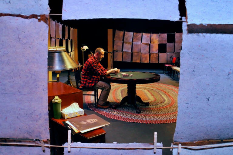 A person is seen through a gap in a row of papers strung on clotheslines. He's seated at a table, looking downwards.