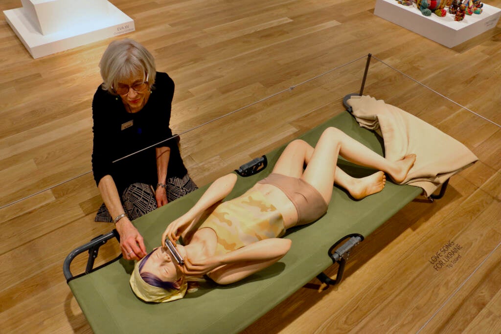 An artist touches the hair on her sculpted figure, a girl lying on a green cot.