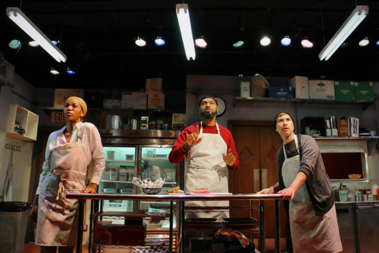 A group of people onstage stand in a kitchen.