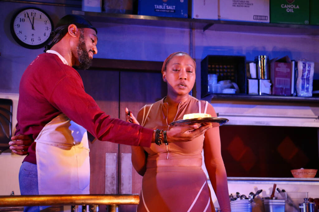 An actor on set presents a woman with a sandwich.