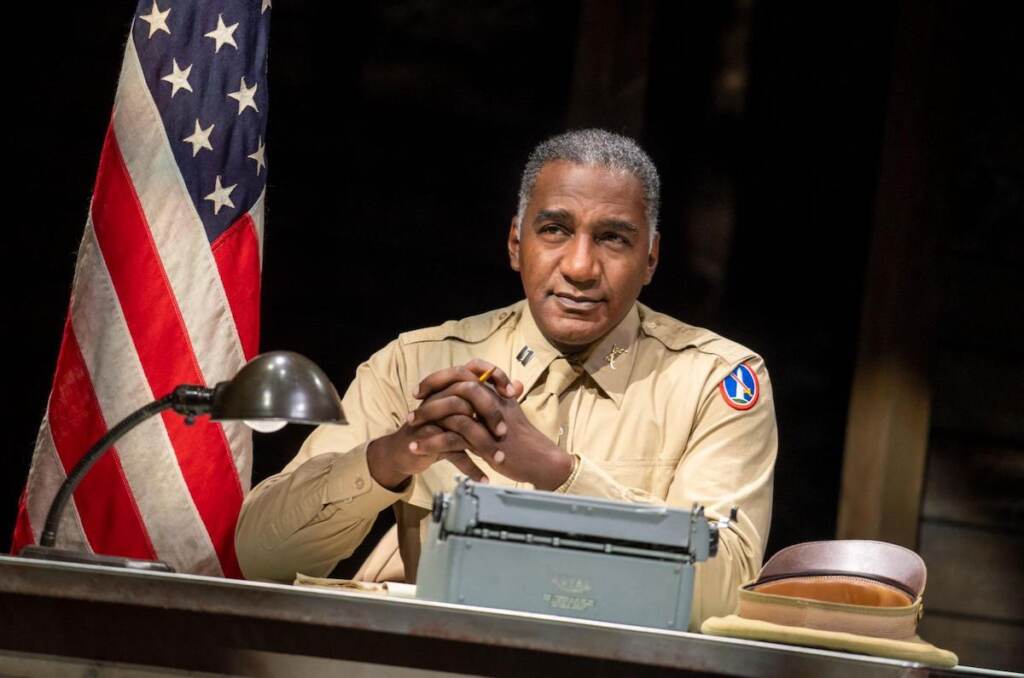 A man in uniform sits at a desk onstage.