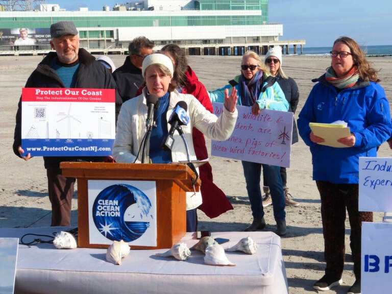 A group of people stand around a podium, some holding signs. The ocean is visible in the background.