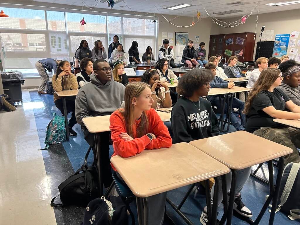 A group of students sit at their desks looking towards the front of the room.