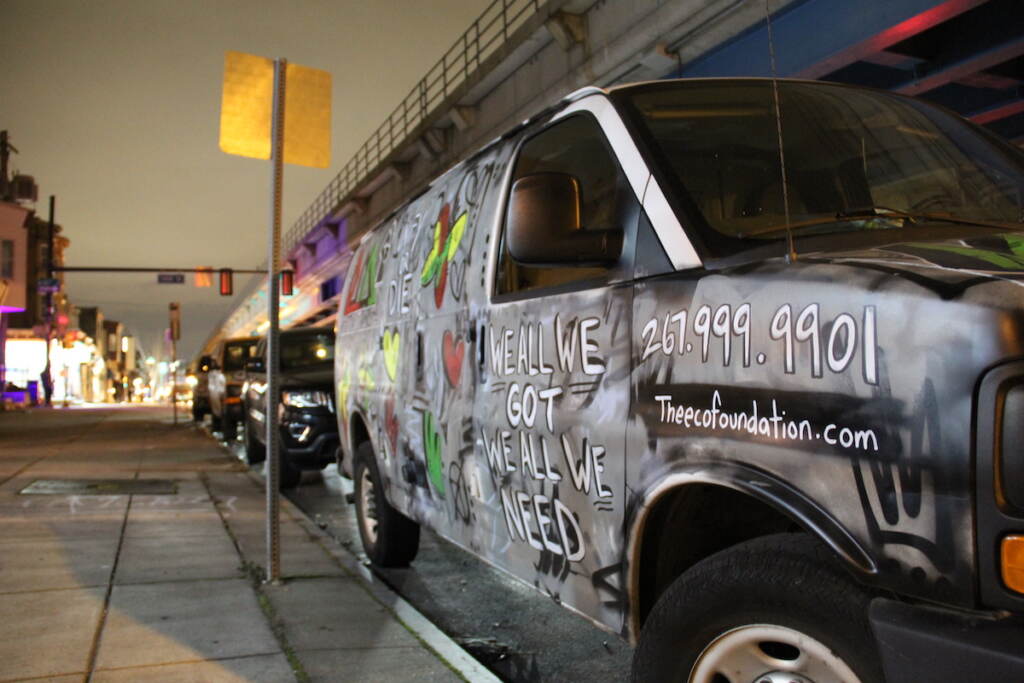 A truck parked by the side of a sidewalk reads "We all we got we all we need." Above the elevated subway tracks are visible.