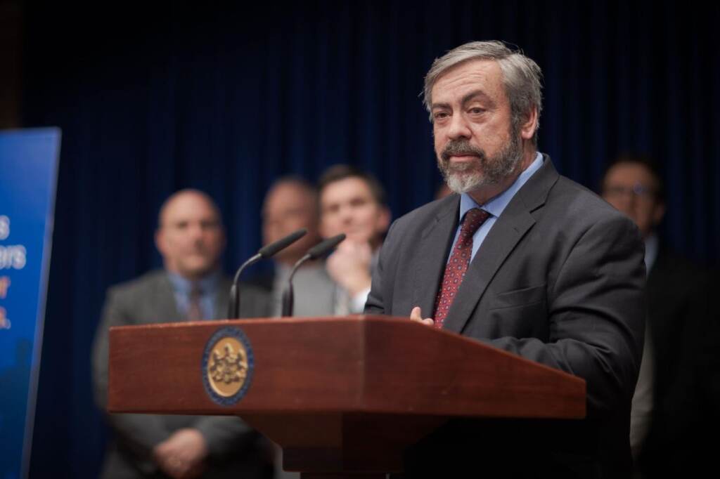 A man stands at a podium. Other people are visible in the background.