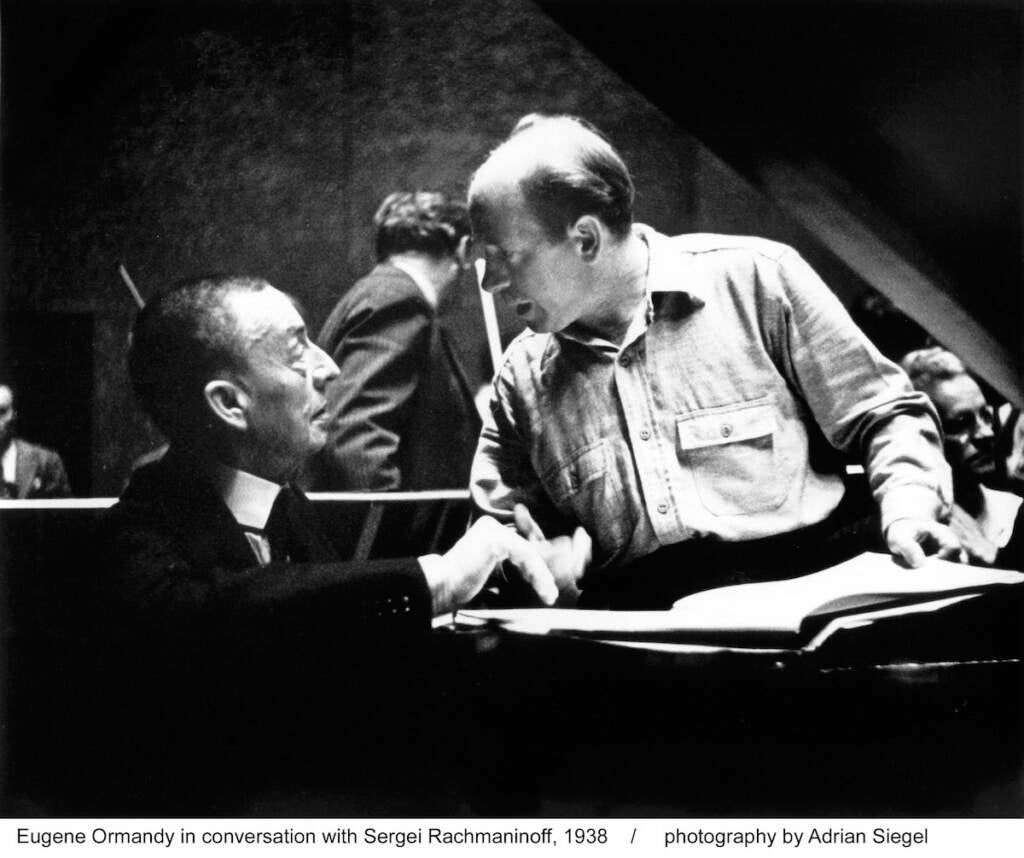 Two men talk at a piano.