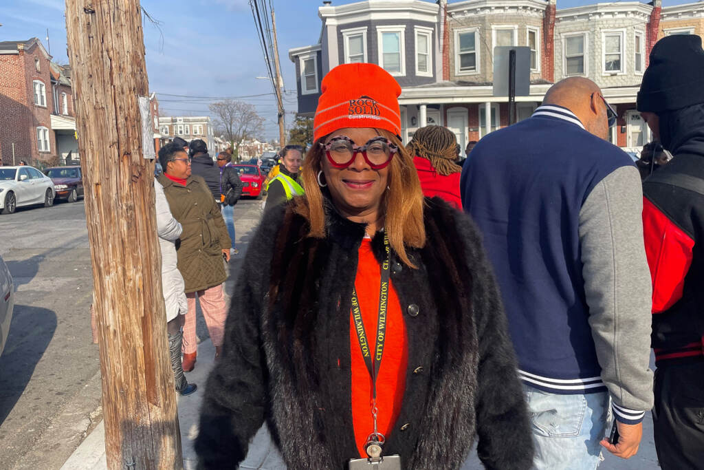 A woman stands in front of the camera, posing for a photo. Groups of people and rowhomes are visible behind her.