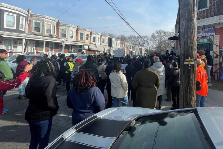 A group of people are gathered on a street lined with rowhomes.