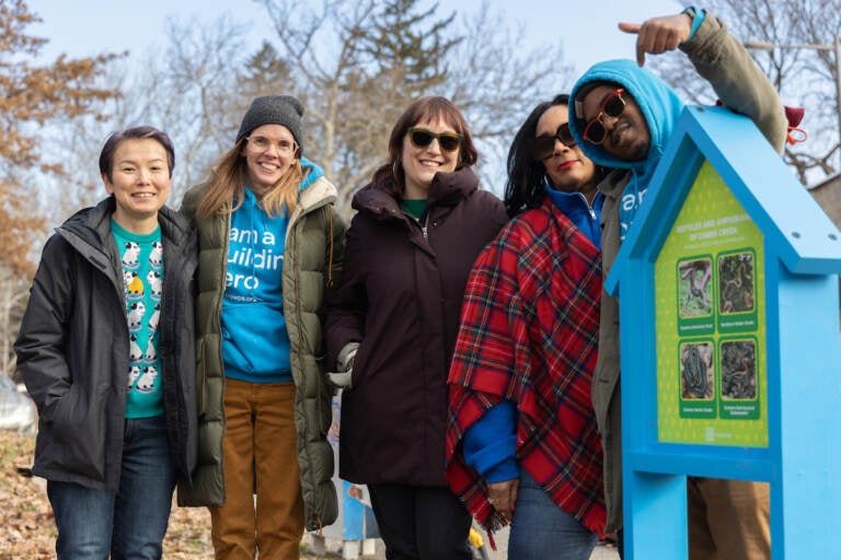 A close-up of a group of people posing for a photo. One person is pointing to a sign.