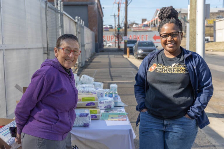 Two people stand, posing for a photo, in front of a table where diapers and other products are laid out.