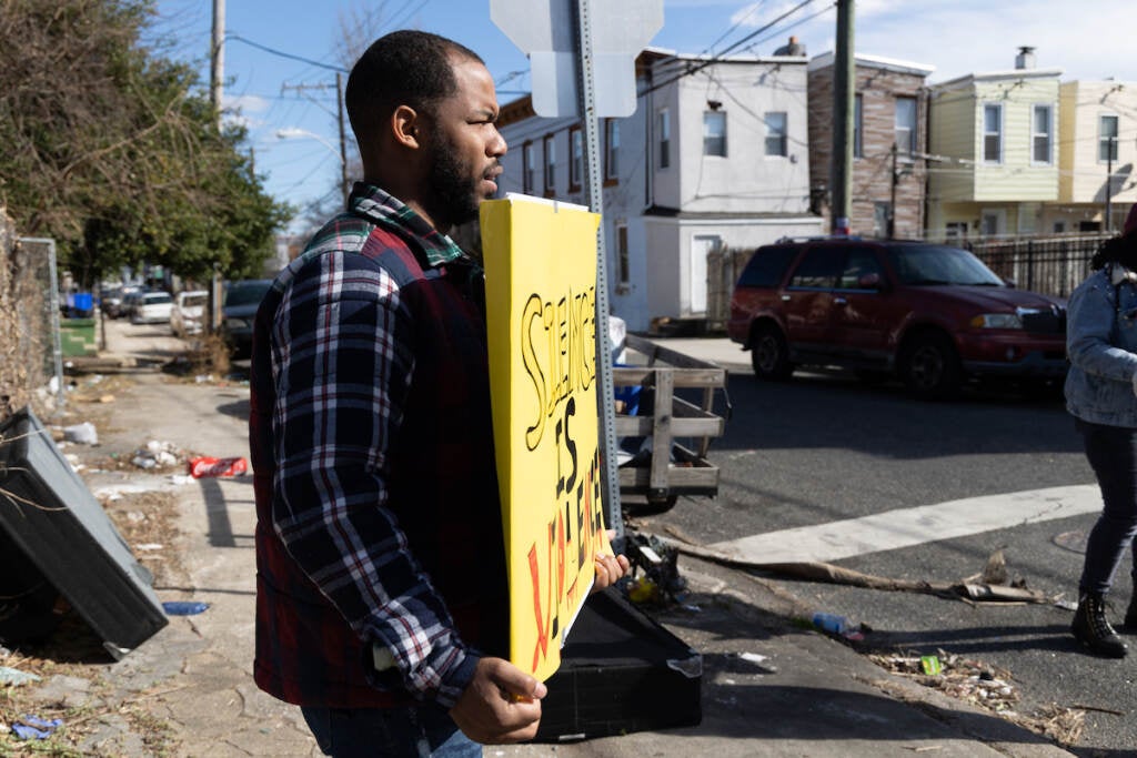 A person holds a sign on a street corner on a sunny day.