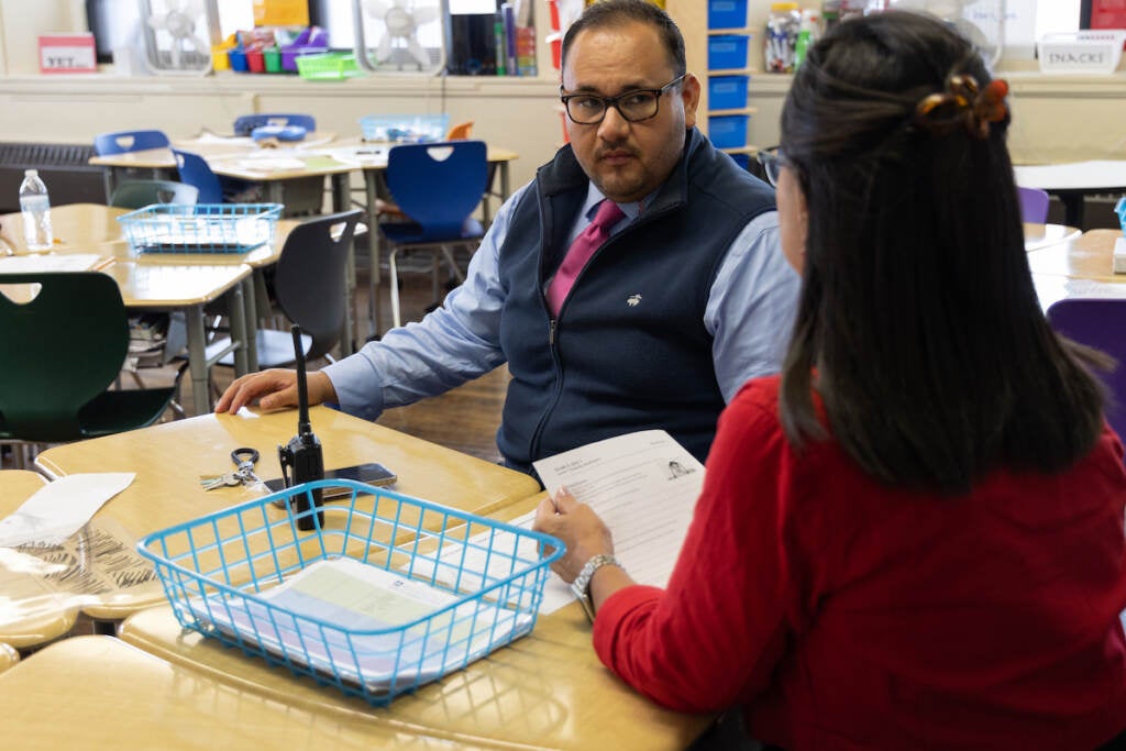 Two people at desks in a classroom, talking.