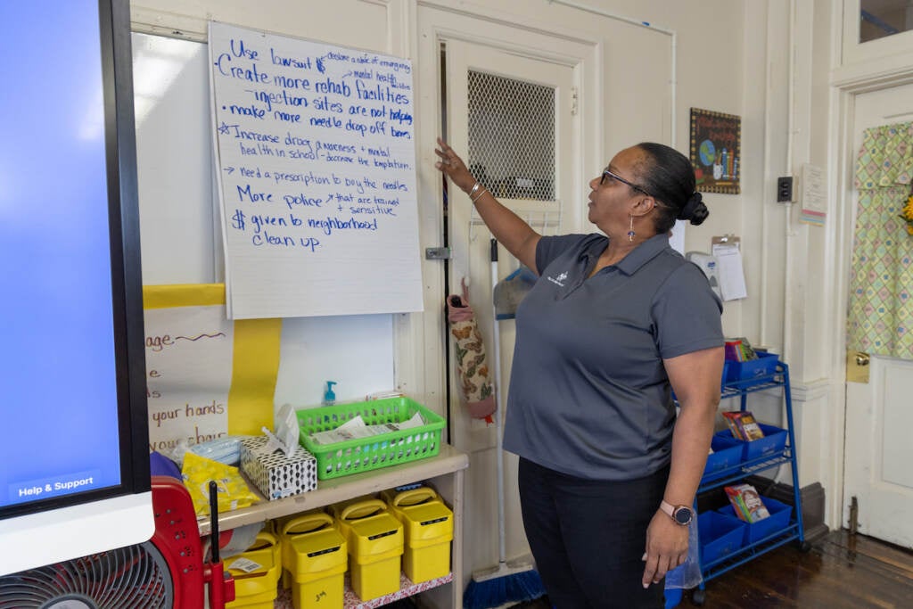 A teacher gestures towards writing on a large piece of paper at the front of a classroom.