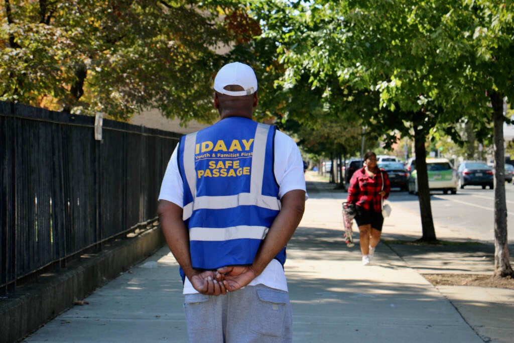 A person wearing a safety vest that reads "IDAAY Safe Passage" stands in the middle of the sidewalk, looking down the street.