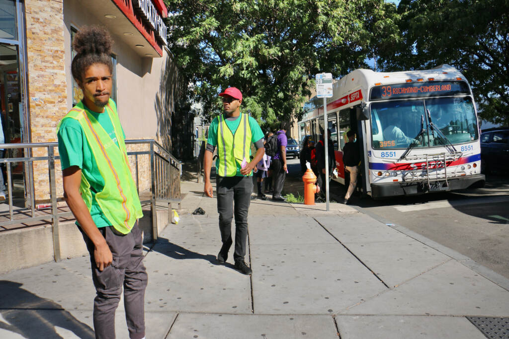 Two people wearing safety vests stand at a street corner. A SEPTA bus and passersby are visible in the background.