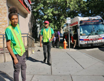 Two people wearing safety vests stand at a street corner. A SEPTA bus and passersby are visible in the background.