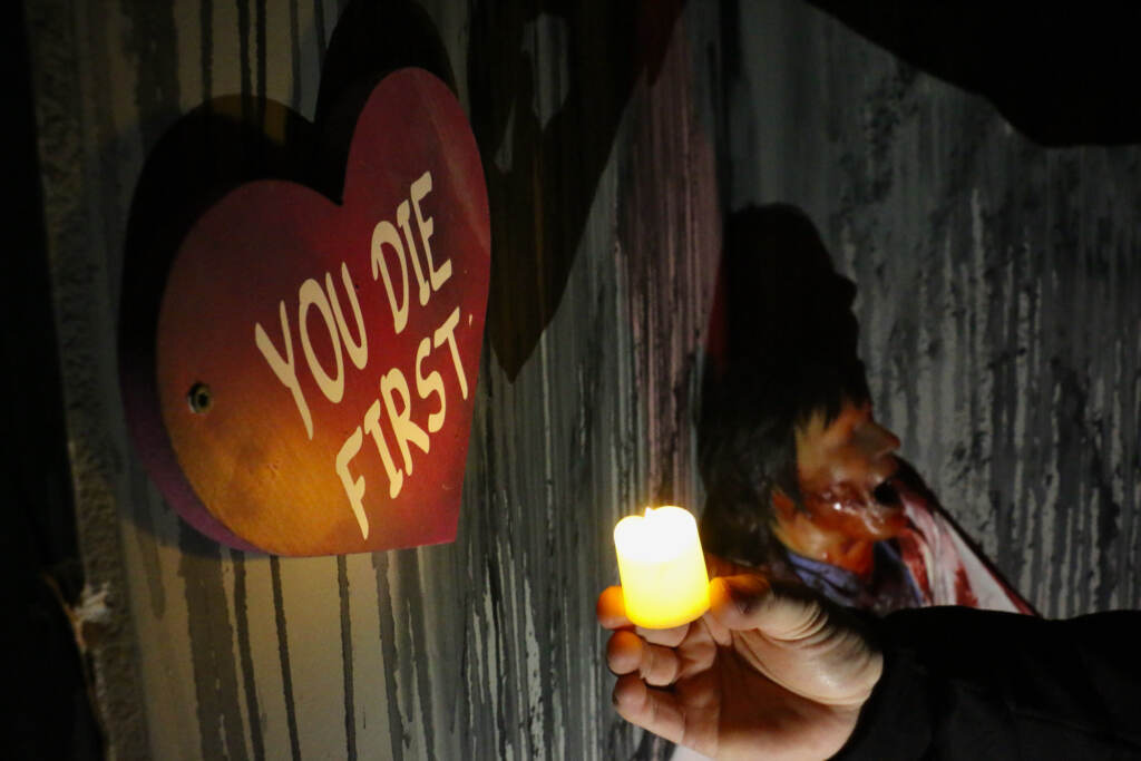 A hand holding a candle lights up a red heart hanging on a wall which reads, "You die first."