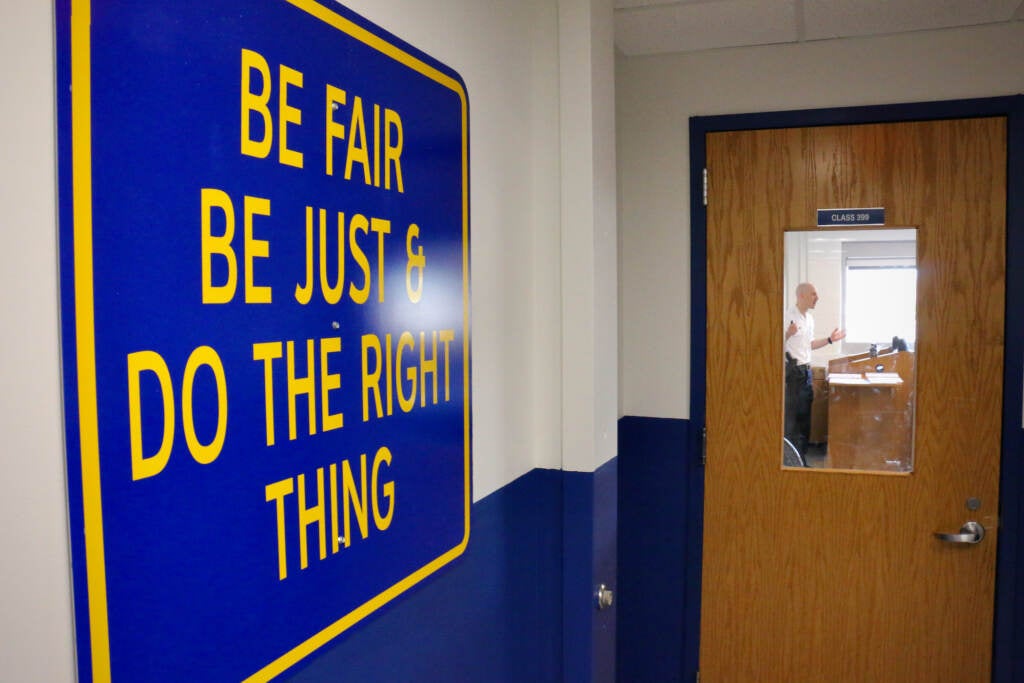 A sign reads "Be Fair, Be Just, & Do the right thing." A person is visible teaching behind a closed door with a window in a classroom.