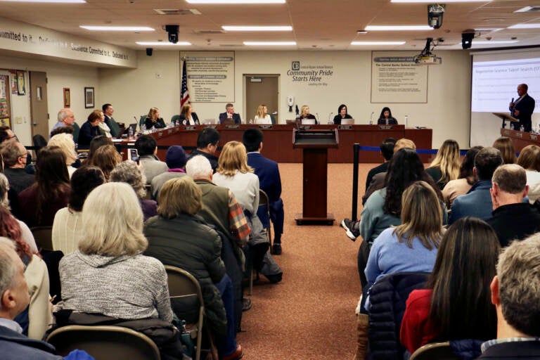 A group of people sit in chairs, and other people sit in a table at the front of the room.