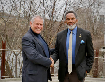 Two men shake hands outdoors while posing for a photo.